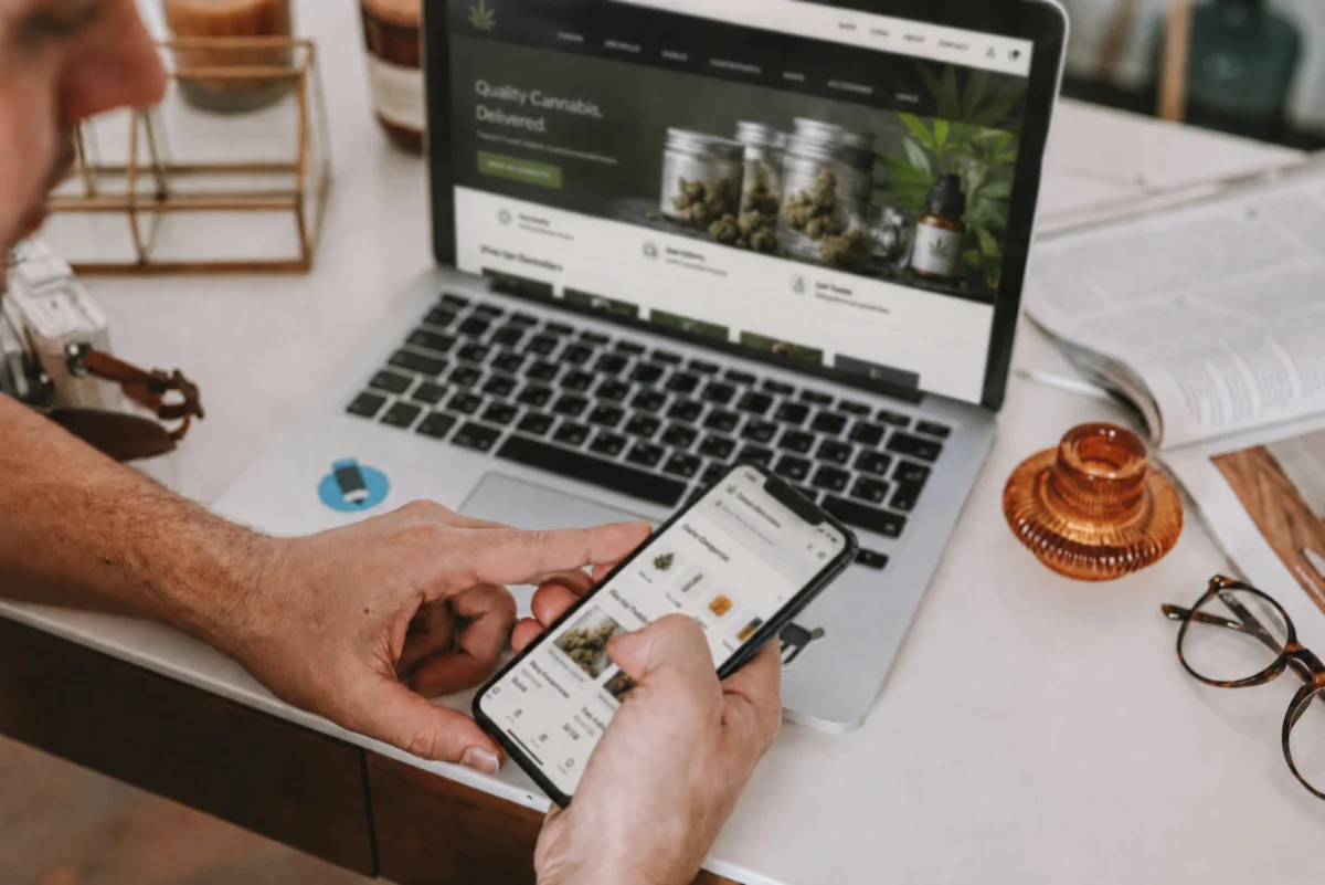 A person browsing a dispensary's online menu on a smartphone while the same cannabis website is open on a laptop at their desk.